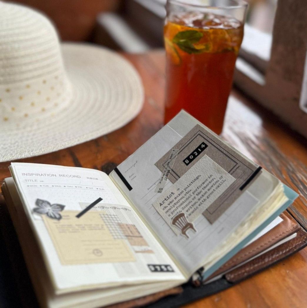 Open notebook with a pen, straw hat, and glass of iced tea on a wooden table.