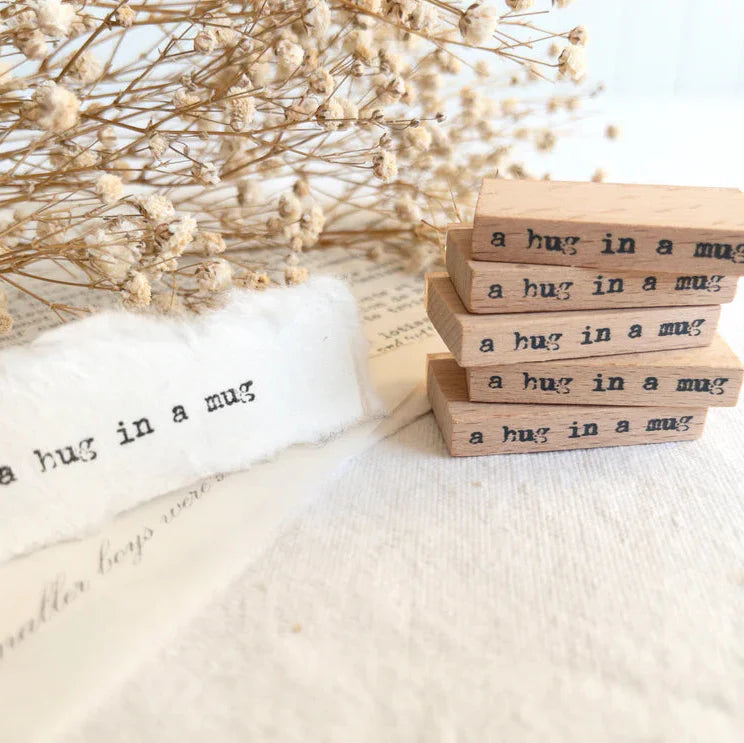 Stack of wooden blocks with 'a hug in a mug' text on a soft surface with dried flowers.