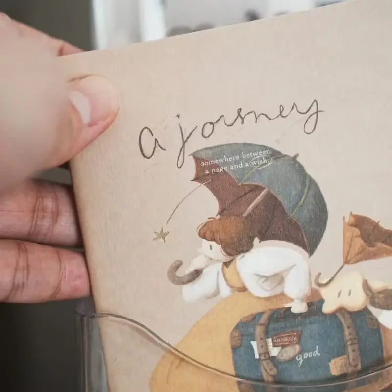 Illustrated notebook with a character holding an umbrella and text 'a journey', held open by a hand.