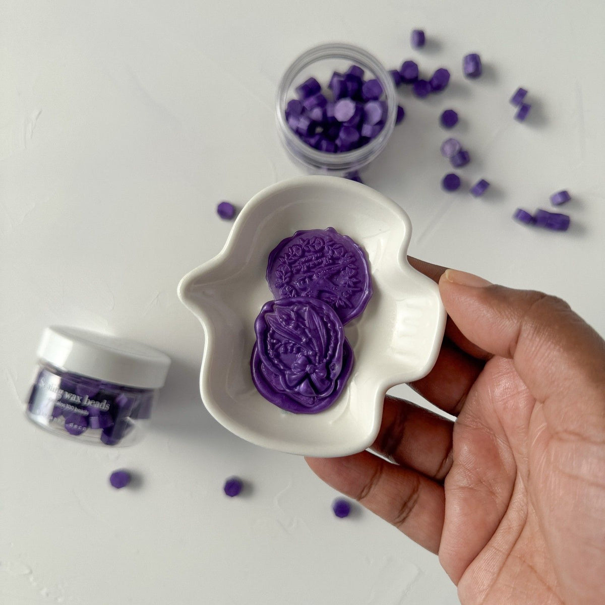Hand holding a white dish of deep twilight-purple Aurora Purple sealing wax beads on a light background