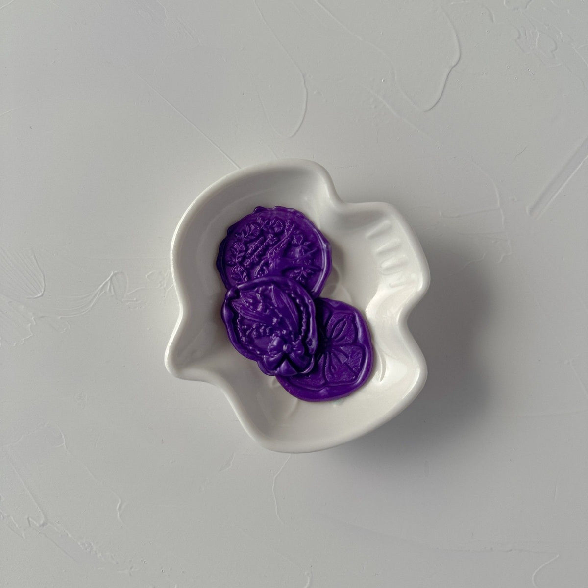 Stamped Aurora Purple wax seal with a floral design displayed in a white dish on a grey background