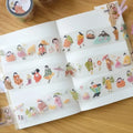 A styled flat-lay of decorative washi tape rolls featuring cute Japanese-style illustrated characters, shown partially unrolled inside an open notebook on a wooden table.