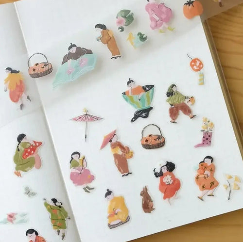 A sheet of small illustrated stickers displayed on a white notebook page, featuring whimsical figures, baskets, flowers, and accessories in soft pastel and bright colors.