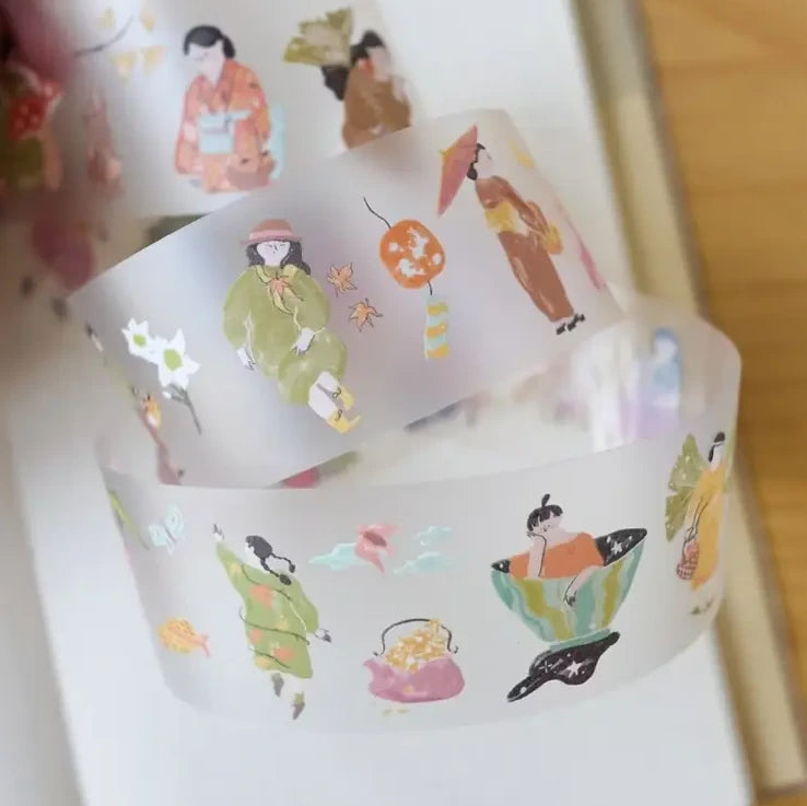 A close-up of a roll of decorative washi tape featuring whimsical Japanese-style illustrations, including figures in traditional clothing, lanterns, fish, and hats, shown coiled on a light surface.