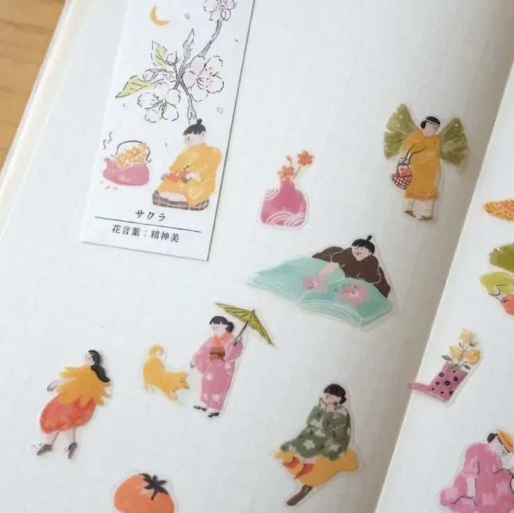 A close-up of a white notebook or journal page decorated with colorful illustrated stickers featuring traditional Japanese figures, flowers, and small objects. One sticker sheet is visible on the left with Japanese text and cherry blossom illustrations.