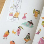 A close-up of a white notebook or journal page decorated with colorful illustrated stickers featuring traditional Japanese figures, flowers, and small objects. One sticker sheet is visible on the left with Japanese text and cherry blossom illustrations.
