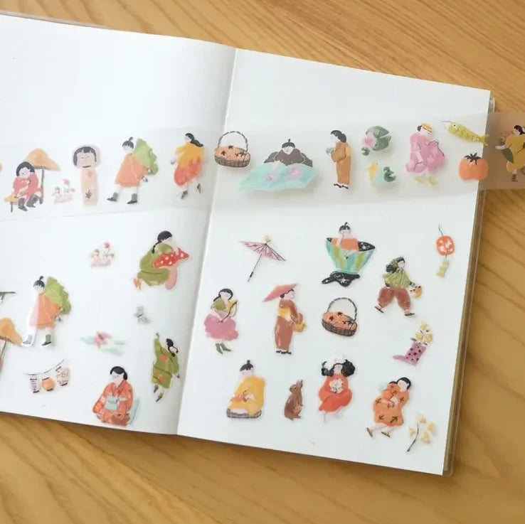 A transparent washi tape roll unrolled and spread across an open notebook, showing small illustrated characters, umbrellas, baskets, flowers, and seasonal motifs on a light wood surface.