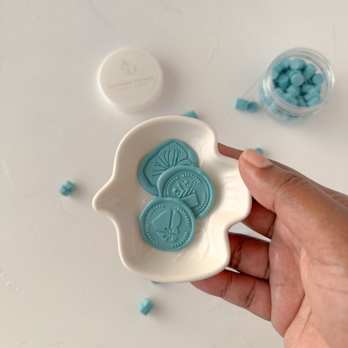 Hand holding a white dish of pastel Baby Blue sealing wax beads against a soft light grey backdrop