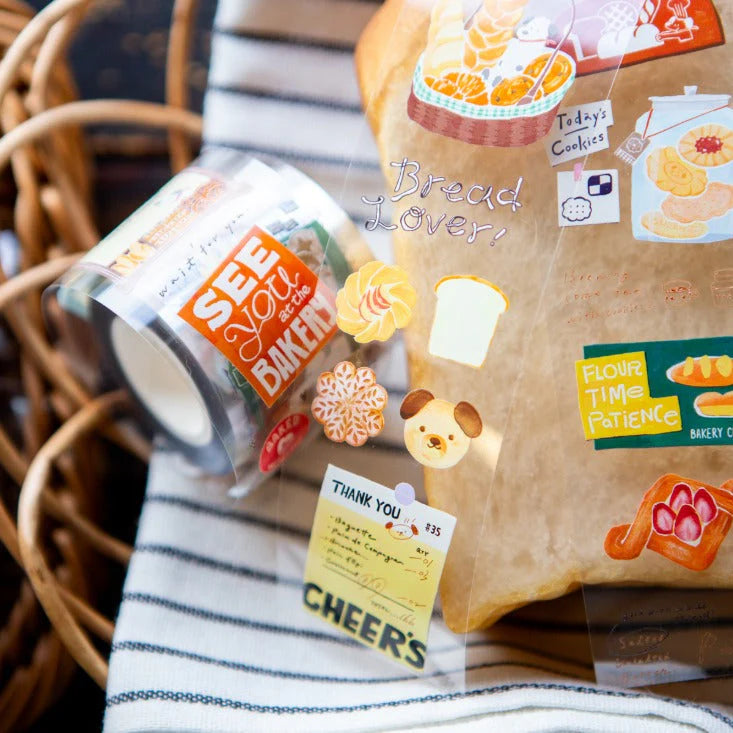 Packaged bread with decorative stickers and a small container labeled 'See Baker' on a striped fabric background.