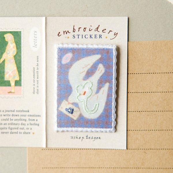 An embroidered sticker displayed on a cream card labeled 'embroidery STICKER' with the signature 'bishop tedyen'. The sticker features a blue background with a white dove holding a green leaf and a small blue bird, set against a styled background with a notebook page on the left and a lined paper sheet on the right.