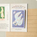 An embroidered sticker displayed on a cream card labeled 'embroidery STICKER' with the signature 'bishop tedyen'. The sticker features a blue background with a white dove holding a green leaf and a small blue bird, set against a styled background with a notebook page on the left and a lined paper sheet on the right.