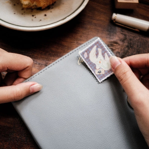 A close-up of a person's hands holding a light gray textured leather notebook or planner cover, with a small square decorative patch attached near the top right. The scene is set on a dark wooden table with a plate and pen blurred in the background.