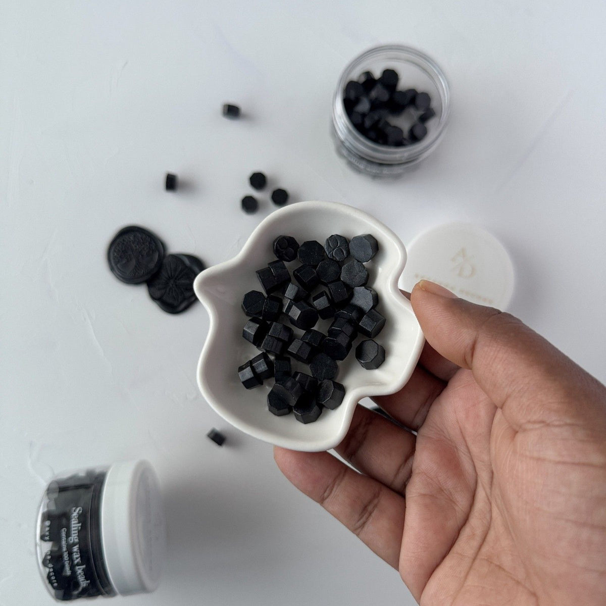Hand holding a white bowl of hexagonal Black Wax Beads sealing wax on a light grey studio background
