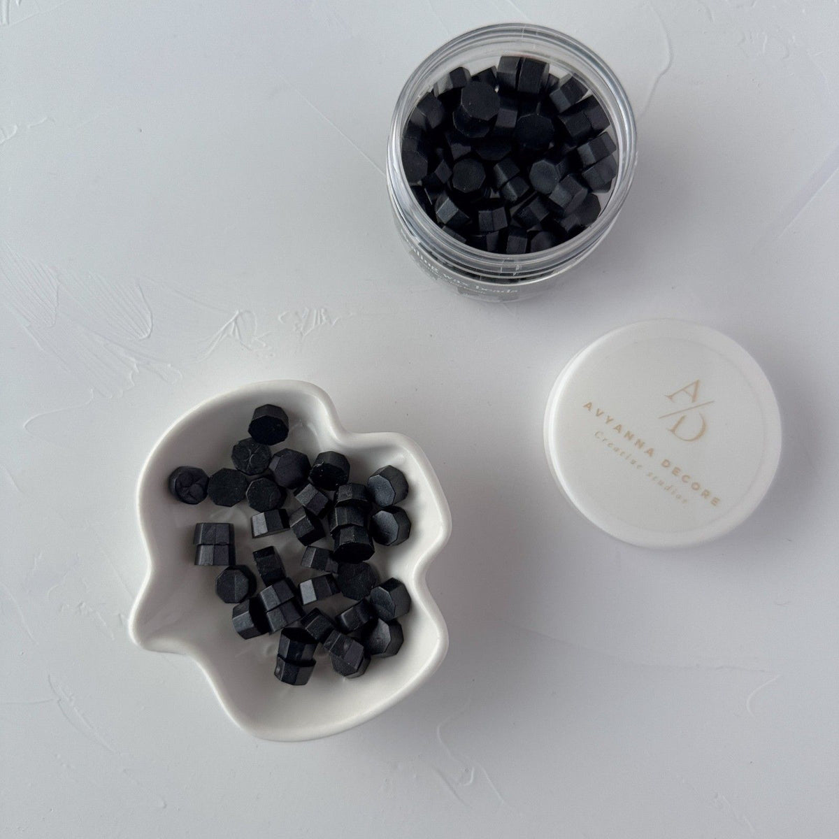 Black hexagonal sealing wax beads in a small dish and jar on a white surface beside a brand card