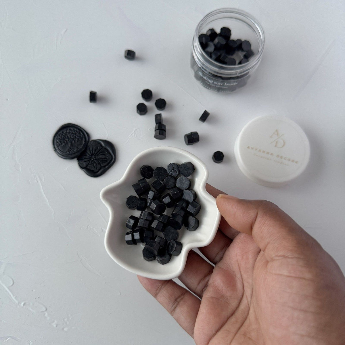 Hand holding a dish of Black Wax Beads with the storage jar and lid behind on a light grey surface