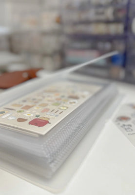 Close-up of a transparent zip organizer pouch holding cute illustrated sticker sheets, placed on a white desk with a softly blurred stationery setup in the background.