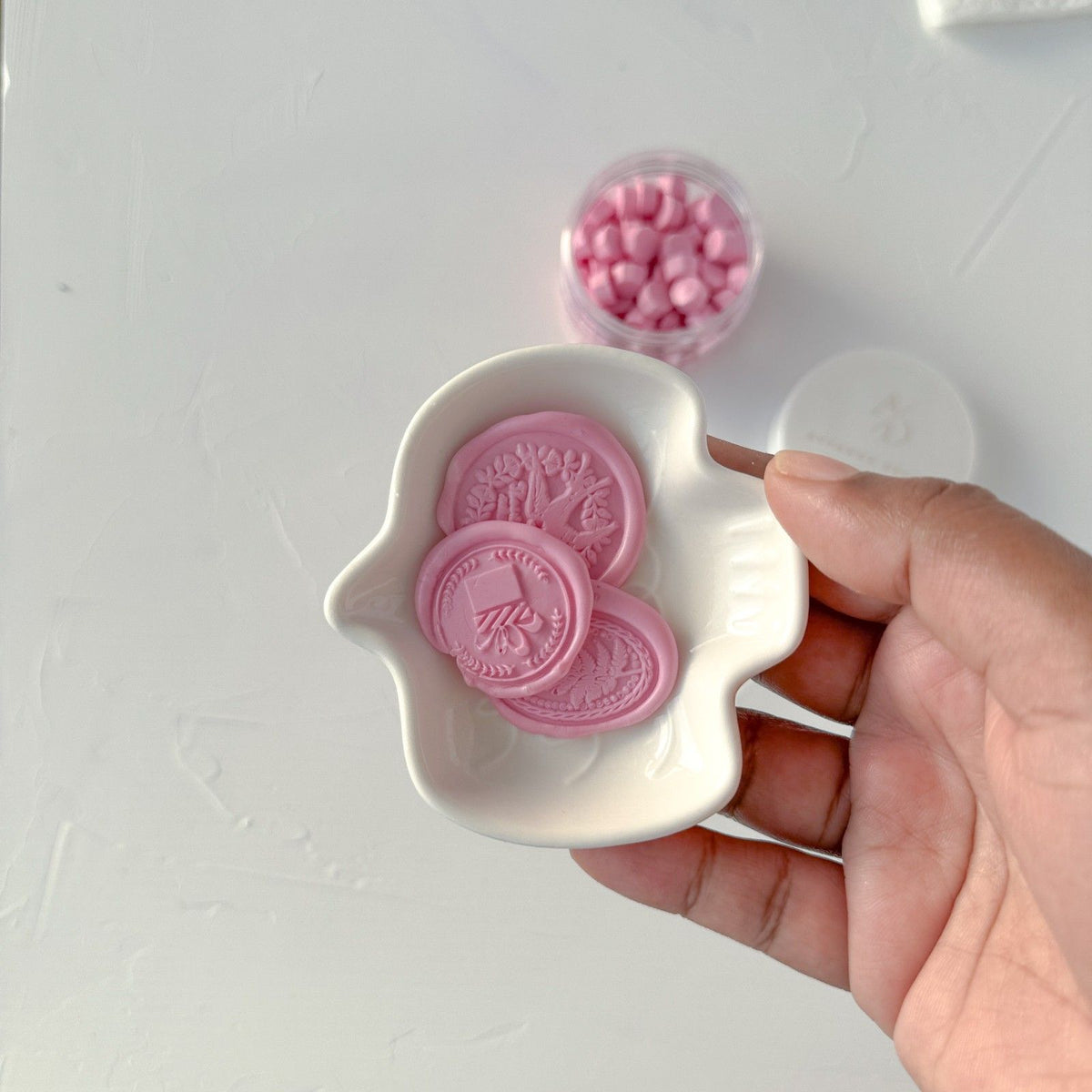Hand holding a white dish of Blossom Wax Beads seals with the container of pink beads behind