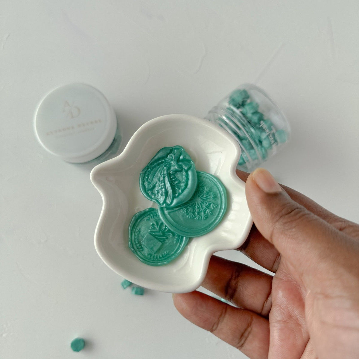 Hand holding a white dish of Blue Lagoon sealing wax beads against a soft light grey studio background