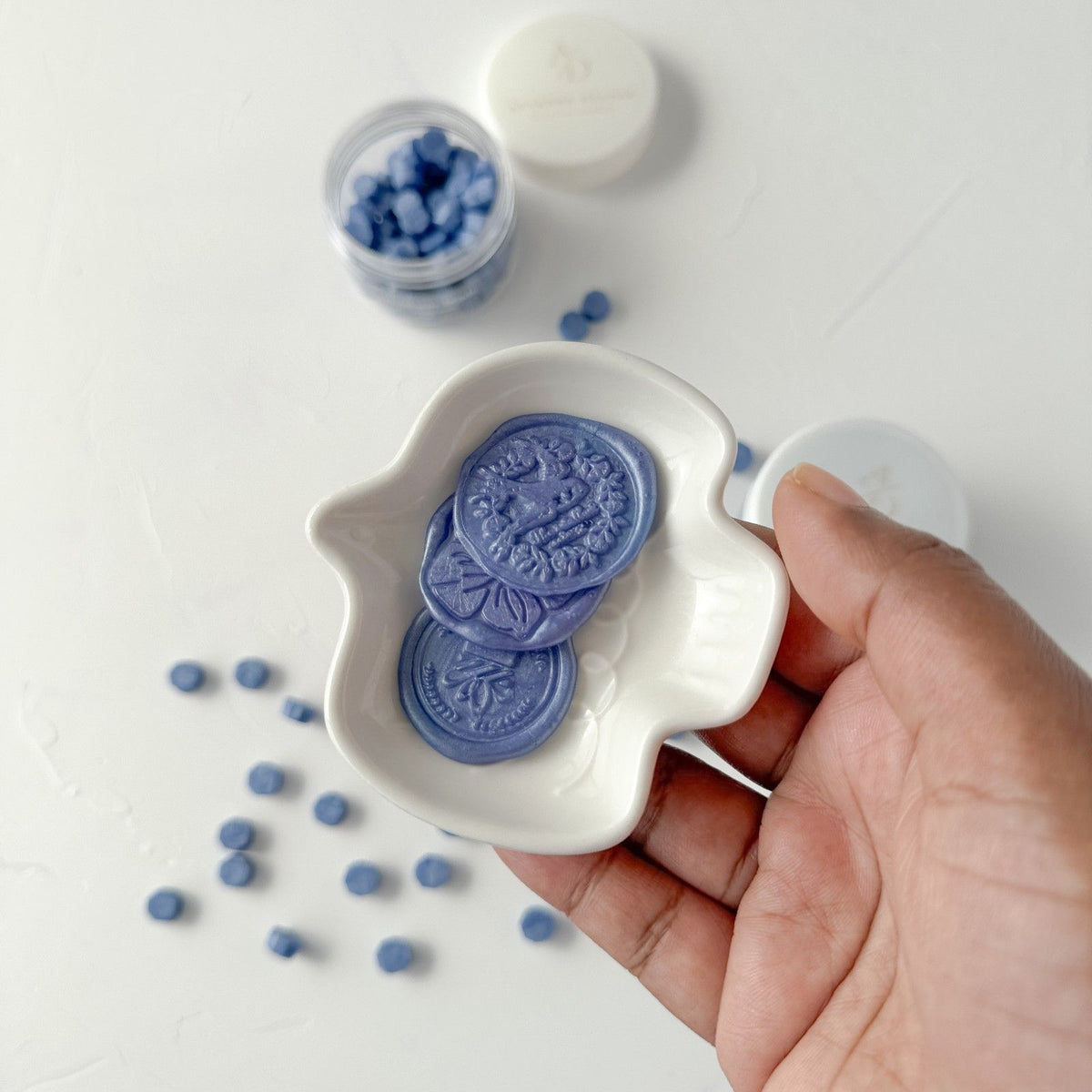 Hand holding a white dish of muted blue Blue Pottery Wax Beads against a light grey studio background