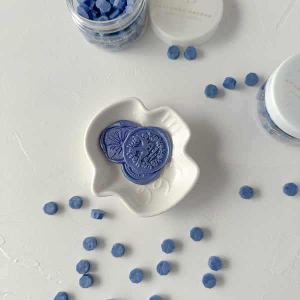 Round Blue Pottery Wax Beads with one crumbled bead showing wax texture, close-up on a white surface