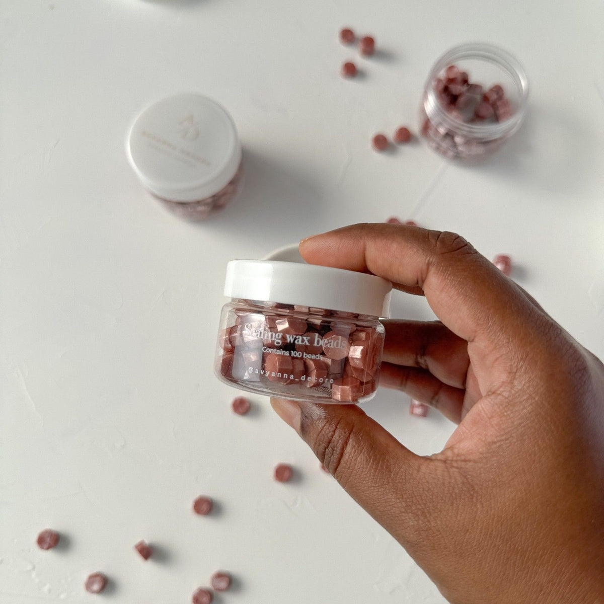 Hand holding the jar of Bottled Grape Wax Beads with its white lid against a soft light grey backdrop