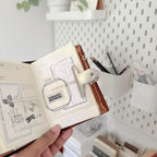 Hand holding a small open book with a metal clamp on top, set against a blurred indoor background.