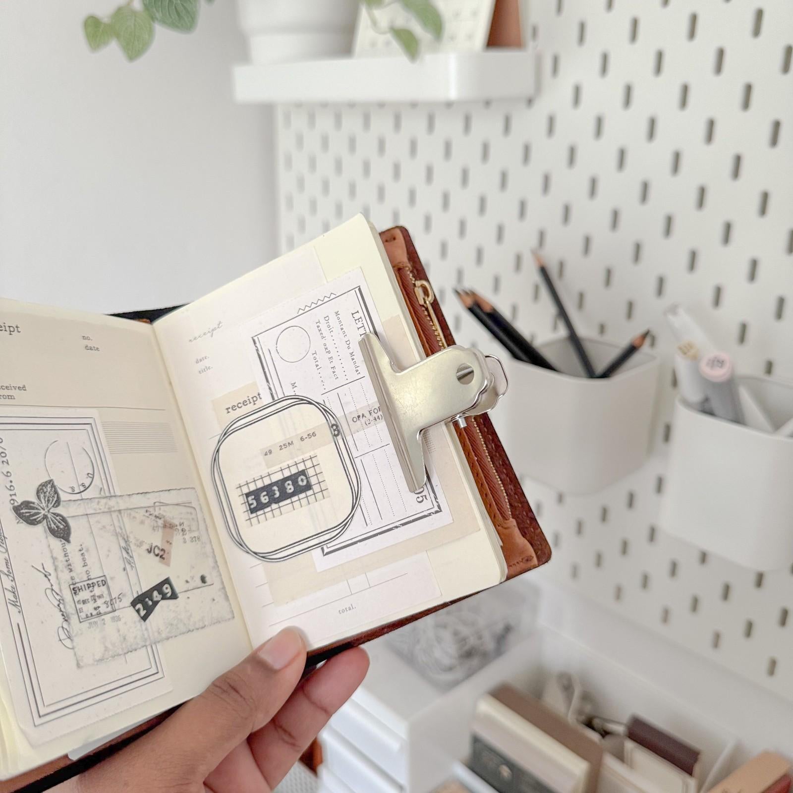 Hand holding a small open book with a metal clamp on top, set against a blurred indoor background.
