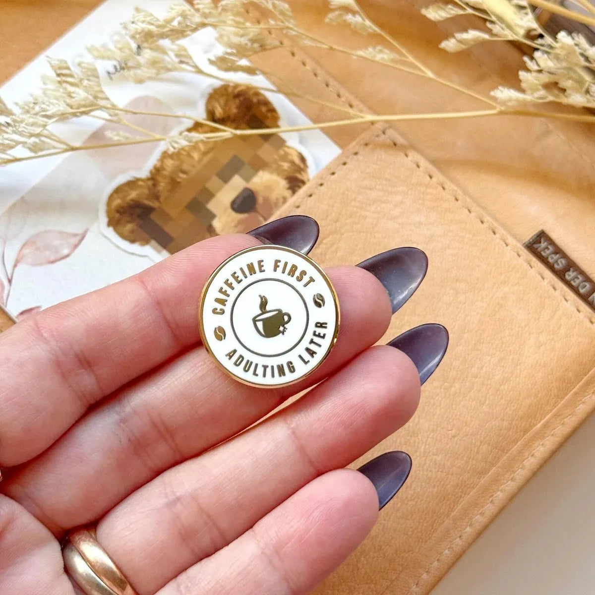 Pin with 'Caffeine First, Adulting Later' text held by a hand with purple nail polish.