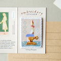 An embroidery sticker featuring a stylized candle in a gold-colored holder, presented on a white backing card labeled 'embroidery STICKER' with handwritten text 'wish tea time' below. The card is styled on a beige notebook page.