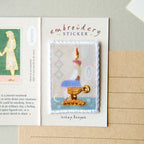 An embroidery sticker featuring a stylized candle in a gold-colored holder, presented on a white backing card labeled 'embroidery STICKER' with handwritten text 'wish tea time' below. The card is styled on a beige notebook page.