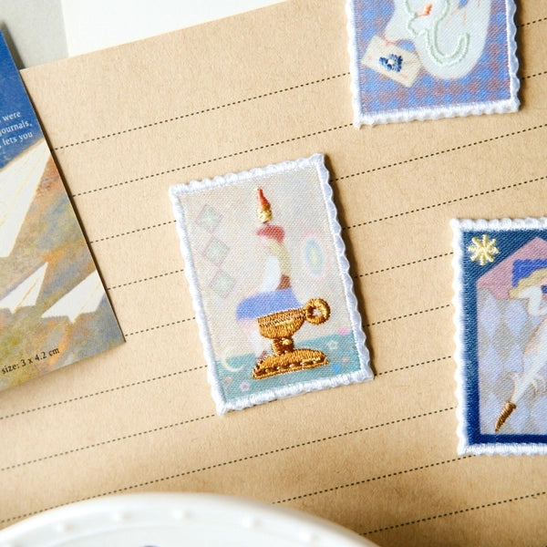 Three small embroidered patch-like stickers with white scalloped edges are arranged on a kraft paper surface. The central patch shows a candle in a gold cup against a pale blue background; the other two patches are partially visible at the top right and right edge and show blue-toned illustrated designs.