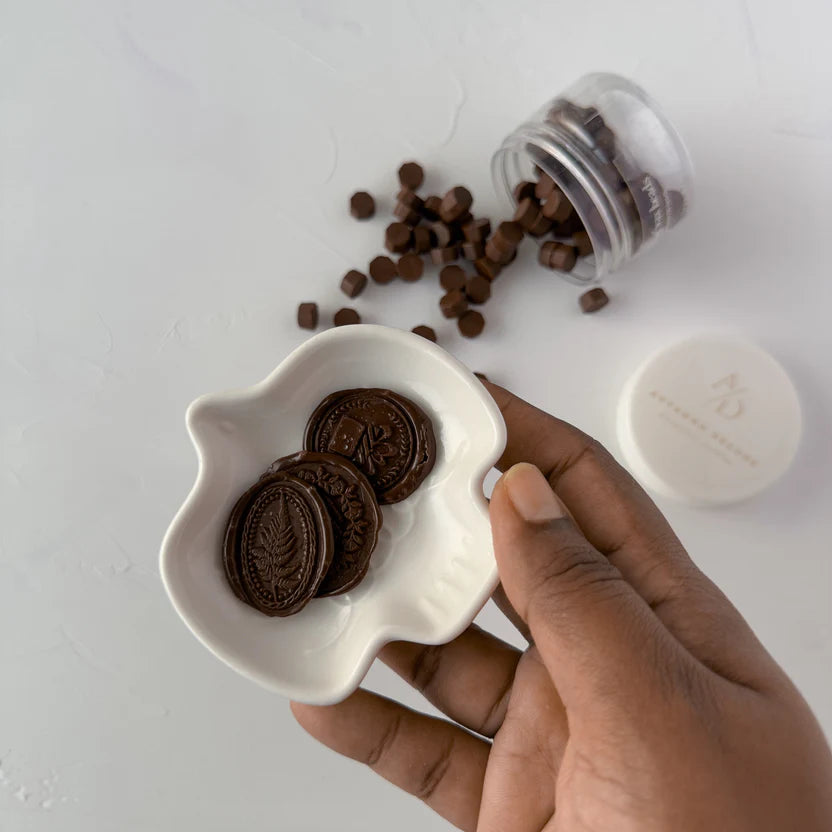 Hand holding a white dish of stamped Chocolate Fudge wax seals surrounded by loose brown beads