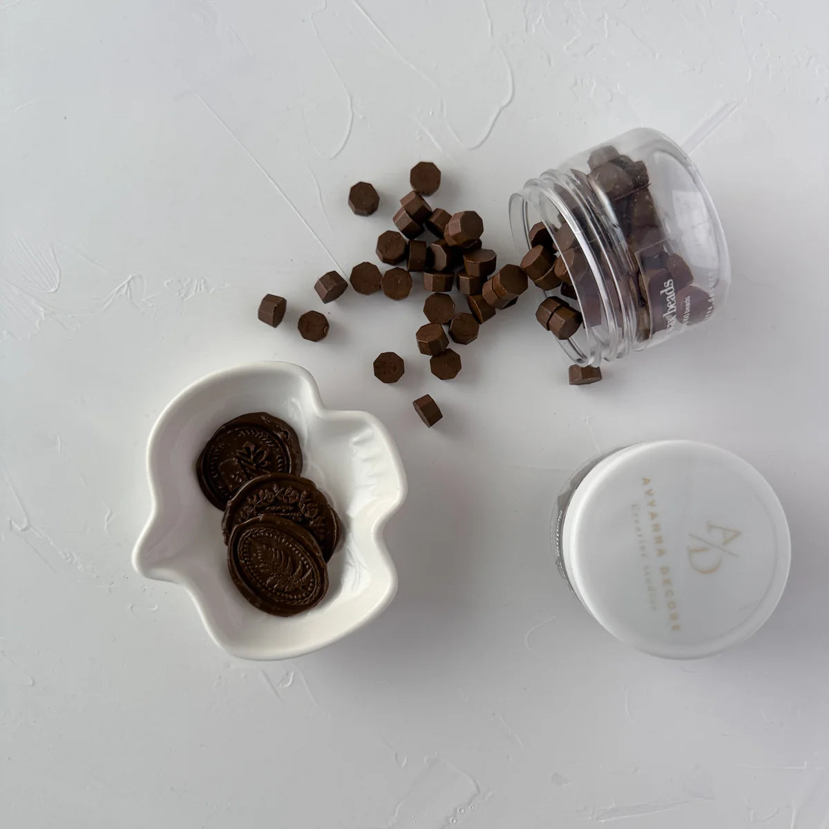 Jar of Chocolate Fudge Wax Beads beside a white dish of stamped seals on a light grey background