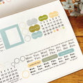 Transparent PET tape with pastel planner stickers including labels, dates, and circular frame designs on wooden desk