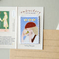 An embroidered sticker card featuring a stitched illustration of a woman with a red beret and yellow flowers on a light blue background, labeled 'embroidery STICKER' with the name 'Ashley Beden' below. The card is displayed on a neutral surface next to a notebook page.