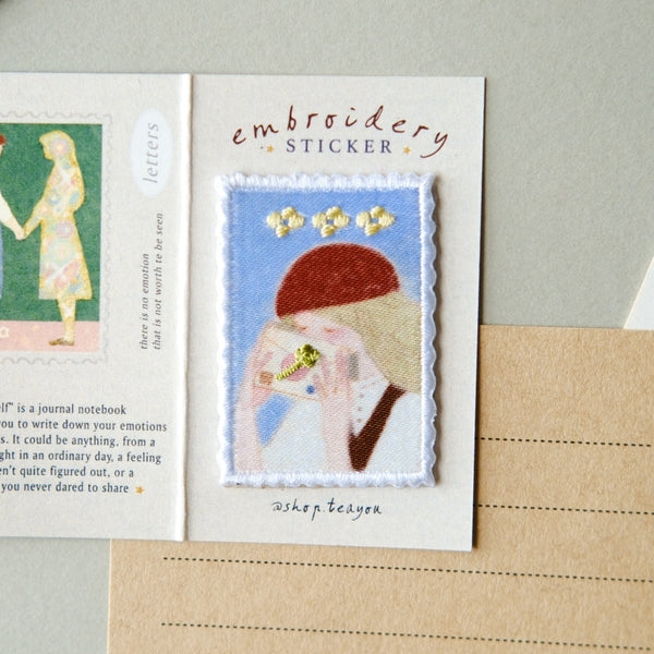 An embroidered sticker card featuring a stitched illustration of a woman with a red beret and yellow flowers on a light blue background, labeled 'embroidery STICKER' with the name 'Ashley Beden' below. The card is displayed on a neutral surface next to a notebook page.