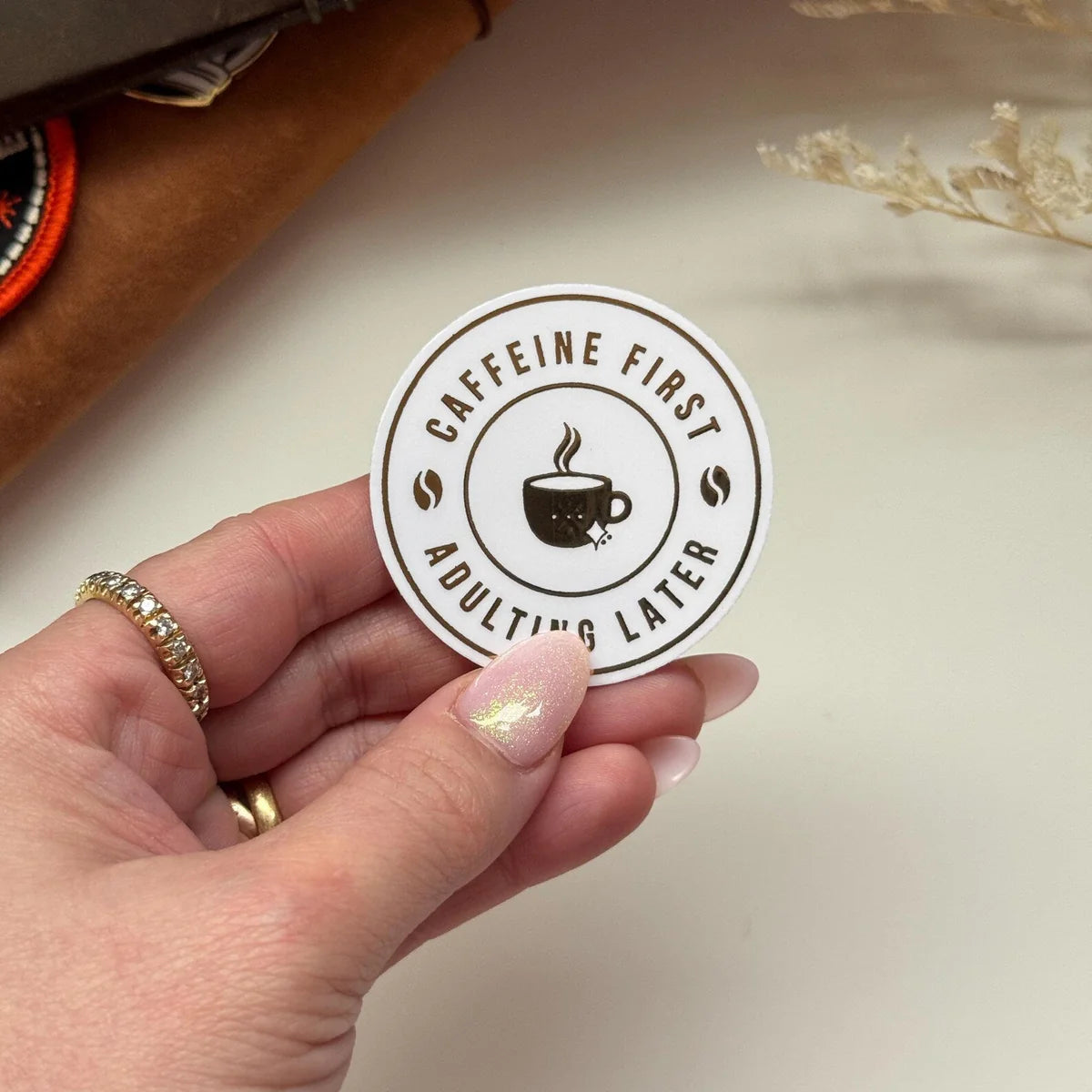 Hand holding a round sticker with 'Caffeine First, Adulting Later' text and coffee cup icon.