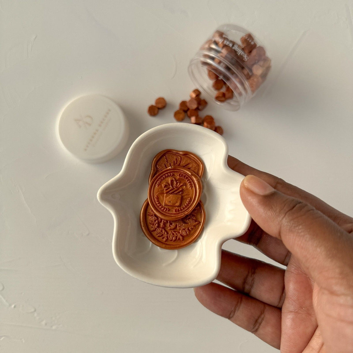 Hand holding a white dish of copper-toned wax seal discs with a container of Copper Wax Beads alongside
