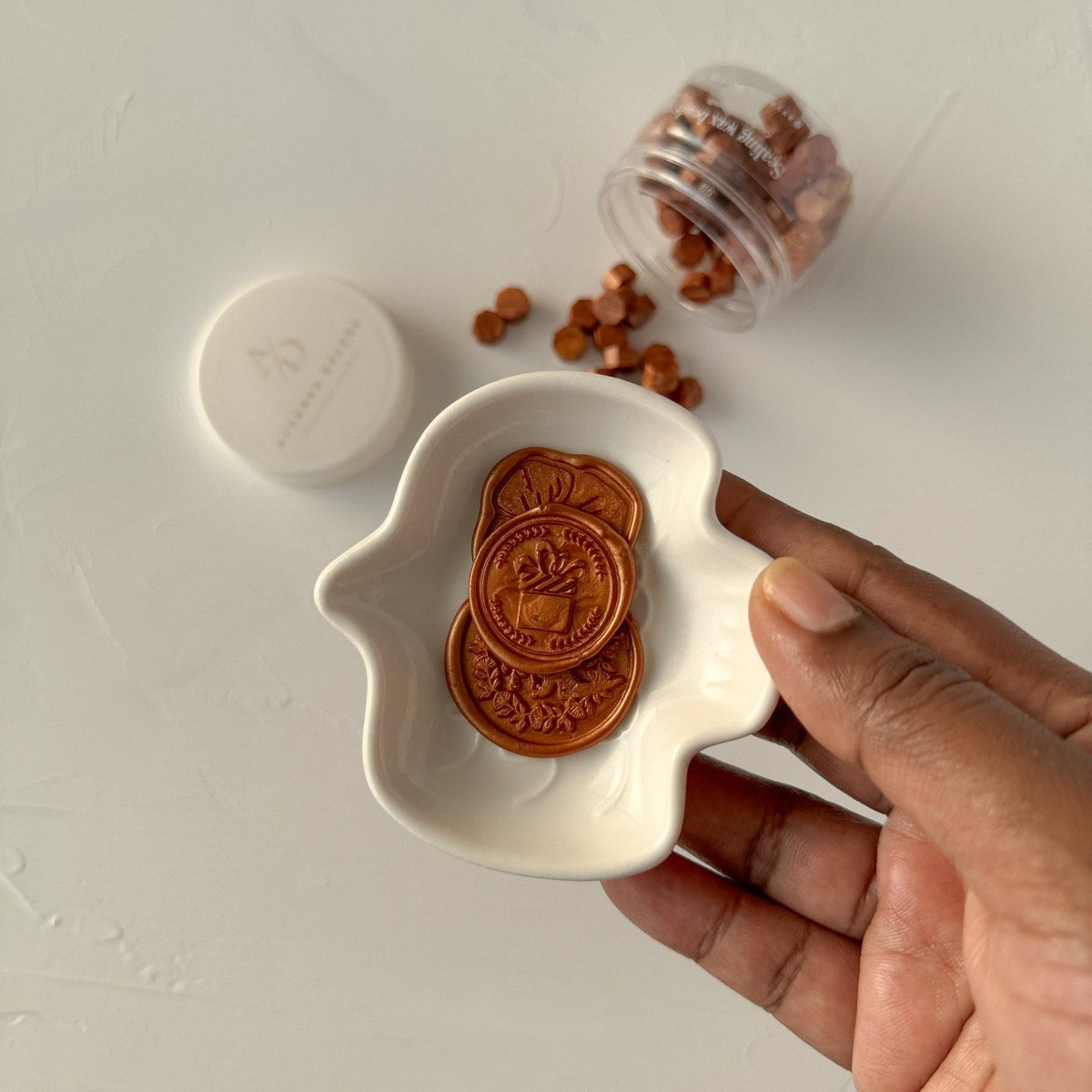 Hand holding a dish of two stamped Copper wax seals with more seals scattered on a light grey backdrop