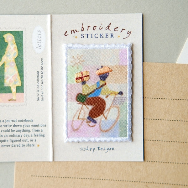 An embroidery sticker card featuring a central rectangular embroidered patch of a person riding a bicycle with a gift box, displayed on a kraft paper envelope.