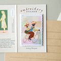 An embroidery sticker card featuring a central rectangular embroidered patch of a person riding a bicycle with a gift box, displayed on a kraft paper envelope.