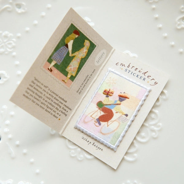 An open handmade card or booklet on a white decorative surface, showing a small embroidered sticker on the right page and printed text/art on the left page. The product appears to be an embroidery sticker or sticker booklet.