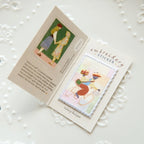 An open handmade card or booklet on a white decorative surface, showing a small embroidered sticker on the right page and printed text/art on the left page. The product appears to be an embroidery sticker or sticker booklet.
