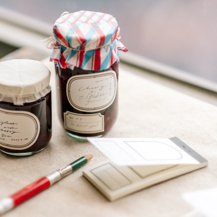 Two jam jars labeled with Ellipse Label Letterpress Book sheets beside a brush and notebook