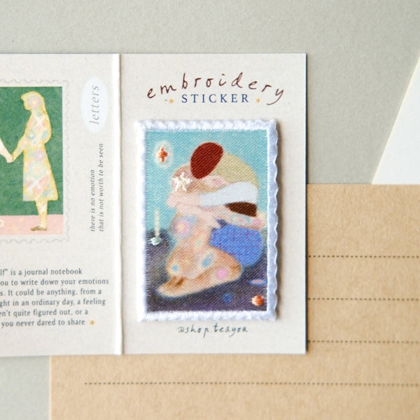 An open white card displaying a small embroidered sticker on the right side and part of another colorful sticker on the left. The right sticker is labeled 'embroidery STICKER' with the name 'shop tea tea' below it, placed on a kraft paper envelope against a light neutral background.