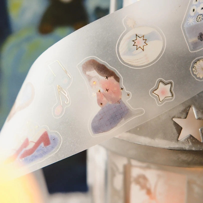 A close-up of a translucent decorative sticker sheet or tape featuring pastel illustrations, including a seated figure, stars, and abstract shapes, draped over a metallic surface with a softly blurred background.
