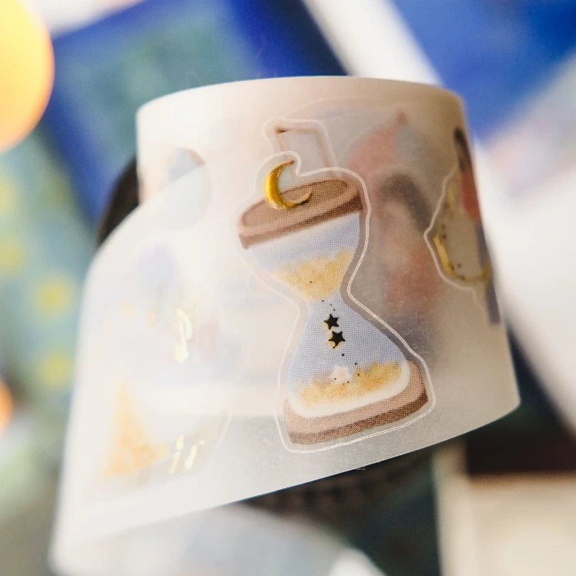 A close-up of a roll of semi-transparent white washi tape featuring whimsical illustrated designs, including a hourglass with sand, stars, and a crescent moon, set against a blurred background.