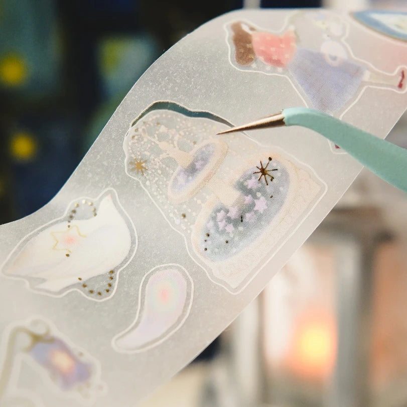 A close-up of a translucent decorative sticker sheet with pastel celestial and animal illustrations, held by a mint green tool against a softly blurred warm-toned background.
