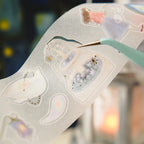 A close-up of a translucent decorative sticker sheet with pastel celestial and animal illustrations, held by a mint green tool against a softly blurred warm-toned background.