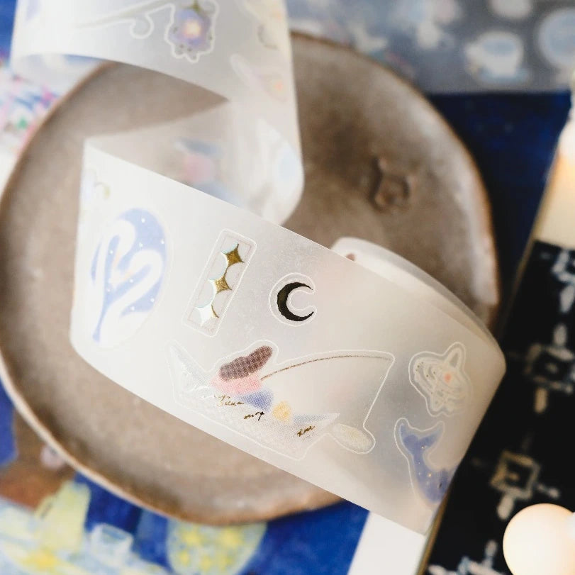 A close-up of a translucent white decorative tape or sticker roll with pastel celestial illustrations, including a crescent moon, stars, and a small flower, draped over a round ceramic dish.